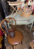 Photo showing wooden bentwood chair with brown cushioned seat, vintage decorative lamp on chair seat, wooden foldable camp stool, and wicker basket on corner desk.