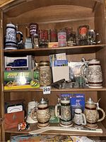 Three wooden shelves filled with assorted boxed diecast vehicles, fire company mugs, steins, small decorative items, and collectible glasses.