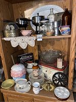 Photo showing two vintage metal fondue pots with forks, assorted glass bottles, small ceramic mugs, metal tins, and large platter on upper shelf with crocheted cloth.