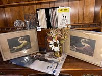 Photo showing two framed vintage-style prints, dog-themed book, fall vase decor, decorative box, clear glassware, and boxes of playbills on a wooden shelf.