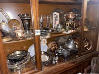 Two wooden glass-door shelves packed with assorted metalware, copper molds, silver trays, pitchers, and glass goblets densely arranged on wooden shelves. Dusty and aged appearance.