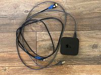 Apple TV box with attached HDMI and power cords displayed on a wooden floor.