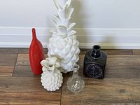 All five vases and decor items grouped on the floor against the wall: red thin nose vase, white pineapple candle holder/vase, clear glass vase, black ceramic nautical box vase, large white pineapple decor item.