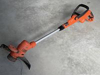 Full view of the weed whacker showing the orange motor housing, black guard near cutting head, straight silver shaft and rear handle.