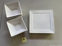 Stack of 5 square plates next to a stack of 4 square bowls on a grey table surface, all white with square shape.