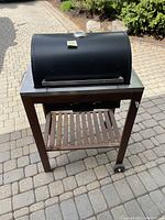 Image of closed charcoal barbecue grill with black curved hinged lid, wooden stand with slatted shelf below and two wheels on one side.