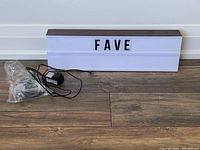 Light message box at front showing the translucent panel with letters spelling 'FAVE', power supply cord and bag of letters placed on the floor.