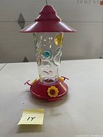 Full view of the hummingbird feeder showing glass container with multicolor circle patterns, red plastic base and lid, and yellow flower feeding ports.