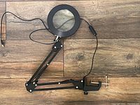 Full view of magnifying loupe arm with clamp, lens, LED ring light, and USB cord on wood floor.