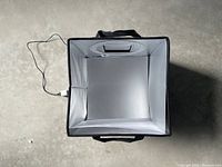 Top-down view of the 12x12 photo lightbox showing interior with reflective silver and white lining, black fabric exterior, and USB cord attached.