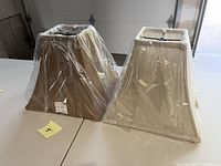 Two rectangular fabric lampshades side by side, one cream and one light brown, both wrapped in clear plastic wrapping on a white surface.