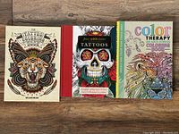 Set of three tattoo coloring books shown side by side on a wooden surface, covers visible.
