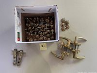 Box containing over 50 solid brass round cabinet knobs and brass-colored door handles with mounting hardware on a white surface