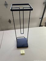 Black metal toilet paper roll holder standing on white table with garage door background; 20 inches tall with square base and 3 vertical rods