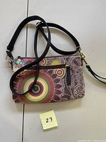 Small wristlet purse shown with colorful circular mandala-style pattern, front zippered pocket, main zippered compartment and detachable black strap with gold-tone clasps.