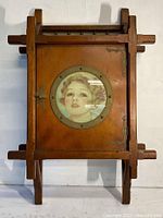 Front view of vintage wooden wall cabinet with porthole showing portrait inside, framed by metal and detailed metal latch and hinges.