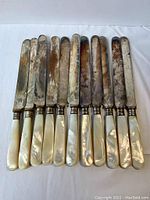 Set of 11 antique butter knives with mother of pearl handles arranged side by side on a white surface showing full length and blade condition.
