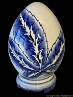 Front view of a large porcelain egg with blue leaf decorative pattern and a matching pedestal base on a black background