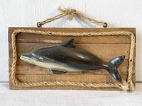 Front view of the carved wooden dolphin mounted on the rectangular wooden plaque with rope border and hanging cord.