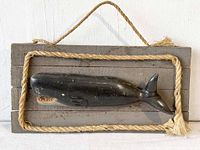 Front view of carved wood whale mounted on gray wooden plaque with rope hanging and border, showing whale length and detailing.