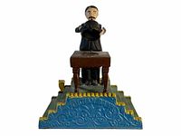 Front view of the vintage cast iron magician bank showing the figure with a mustache holding a black top hat, standing behind a table on an ornate blue base with yellow accents.