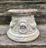 Wide front view of the vintage cement garden pedestal showing lion head with ring and acanthus garland decorations on a weathered surface.