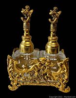 Photo showing front view of both clear square glass perfume bottles with gold plated cherub toppers and ornate holder featuring cherub and floral details.
