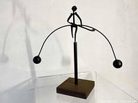 Front view of kinetic metal sculpture showing person balancing on curved rod with spherical weights, mounted on wood base.