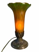 Front view of vintage glass lamp lit, showing green and amber glass shade and metal ornate base.