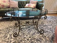 Side and angle view of the oval metal framed coffee table with clear glass top on a patterned rug in front of a green couch.