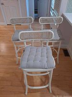 Three white counter stools arranged closely; visible rattan woven backs and light gray tufted cushions with wooden curved legs on hardwood floor.
