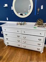 Front view of white wooden dresser showing eight drawers, photographed against blue wall.