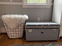 Photo showing wire hamper with white fabric liner and faux shag throw on top, next to gray storage trunk.