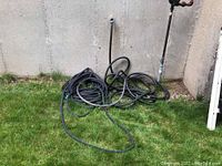 Two black garden hoses coiled near an outdoor water spigot against a wall. One hose is thicker and black nylon-cased, identified as Zero-G brand. The other hose is thinner and plain black rubber/plastic hose.