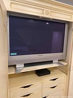 Full view of the Panasonic plasma TV in a silver/grey frame with speaker grille and pedestal stand, placed inside a cream entertainment cabinet.