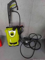 Photo of green and black Sunjoe SPX3000 power washer with attached spray gun and hose alongside Rainbow Lifegard submersible pump on floor against grey wall.