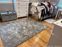 Photo showing larger gray plush area rug on a hardwood floor placed near white dresser and bed with laundry on it.