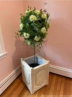 Faux hydrangea topiary with cream flower clusters and green leaves standing in a distressed white resin planter with raised panels, placed in corner against pink walls and wooden baseboards.