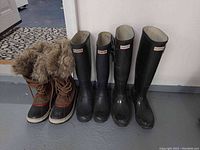 Three pairs of women's boots aligned on the floor: two pairs of black Hunter rain boots and one pair of brown Sorel winter boots with fur lining and laces