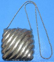 Photo of closed silver tone metal handbag with curved waveform shell design and braided chain strap against blue background.