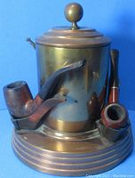 Front view of the copper and brass pipe tobacco humidor with three pipes resting in the holders showing pipe bowls and stem shapes.