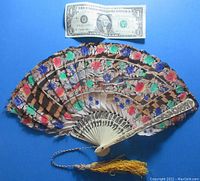 Feather fan opened flat next to a US dollar for scale, showing colorful painted feathers and carved bone ribs with floral patterns.