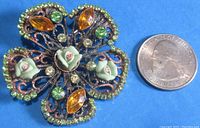 Front view of floral pin showing rhinestones, ceramic rose buds, and green and copper metalwork beside a US quarter for scale.