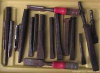 Photo showing multiple metal punches, chisels, and hammers with some red handle grips, arranged flat on a yellow surface.