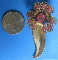 Photo showing the pin next to a quarter coin for size reference (approximately 2.5 inches). The floral design is gold-tone metal with pink rhinestones, featuring a detailed textured leaf dangling from the flower.