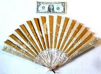 Full view of the open fan with a US dollar bill for size reference, showing the alternating beige and off-white fabric with silver embroidered bird and branches, and abalone ribs.