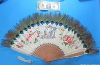 Full view of the fan showing white feathers mounted on sandalwood ribs, peacock feather eyes, hand-painted Oriental couple and floral designs, with a dollar bill for scale.