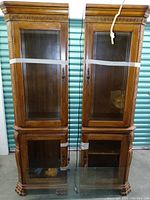 Full view of two wood china hutch end units side by side, showing glass display doors, crown molding, taped together for stability, cords for lighting hanging down.