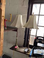 Pair of matching brass lamps standing on a small table near a window, white bell-shaped fabric shades, brass bases with decorative elements, pull chains visible.