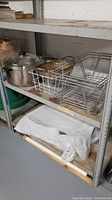 Photo showing shelving with metal cooking pot on left, metal wire basket behind it and three metal chafing trays stacked on right, all with surface rust and in used condition.